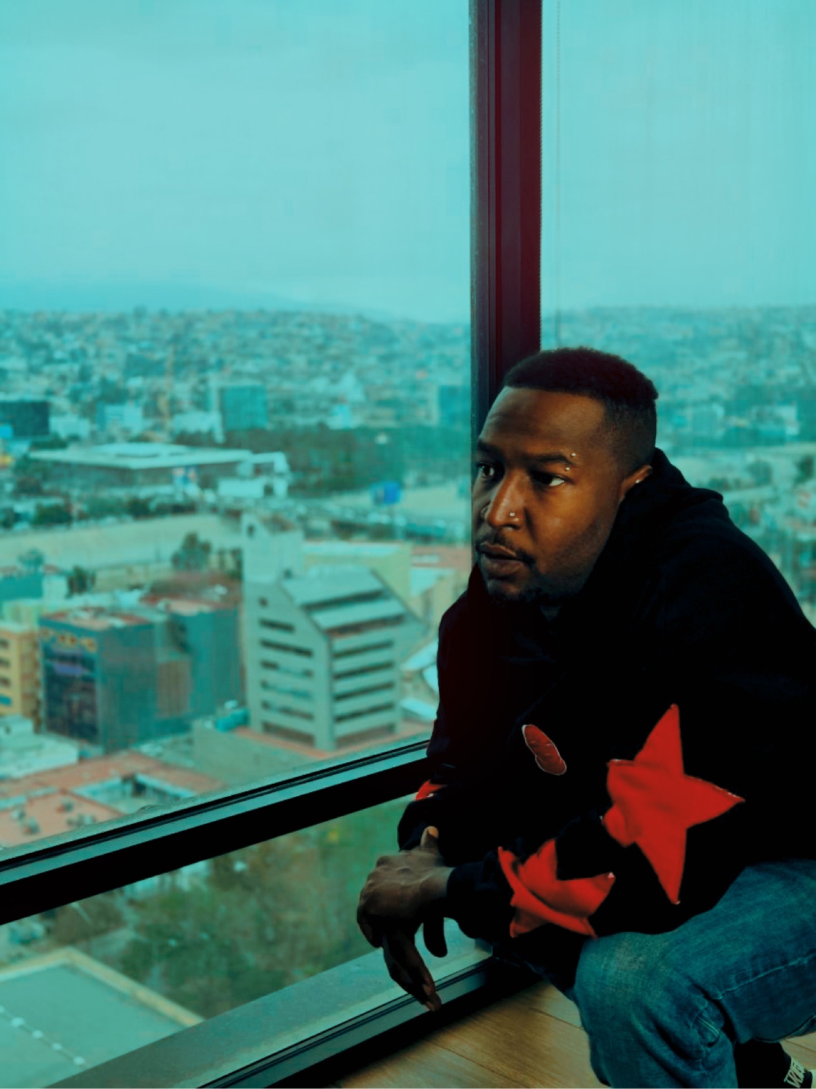 Cortez Devinci in a black hoodie with red stars, seated by a tall window overlooking the city