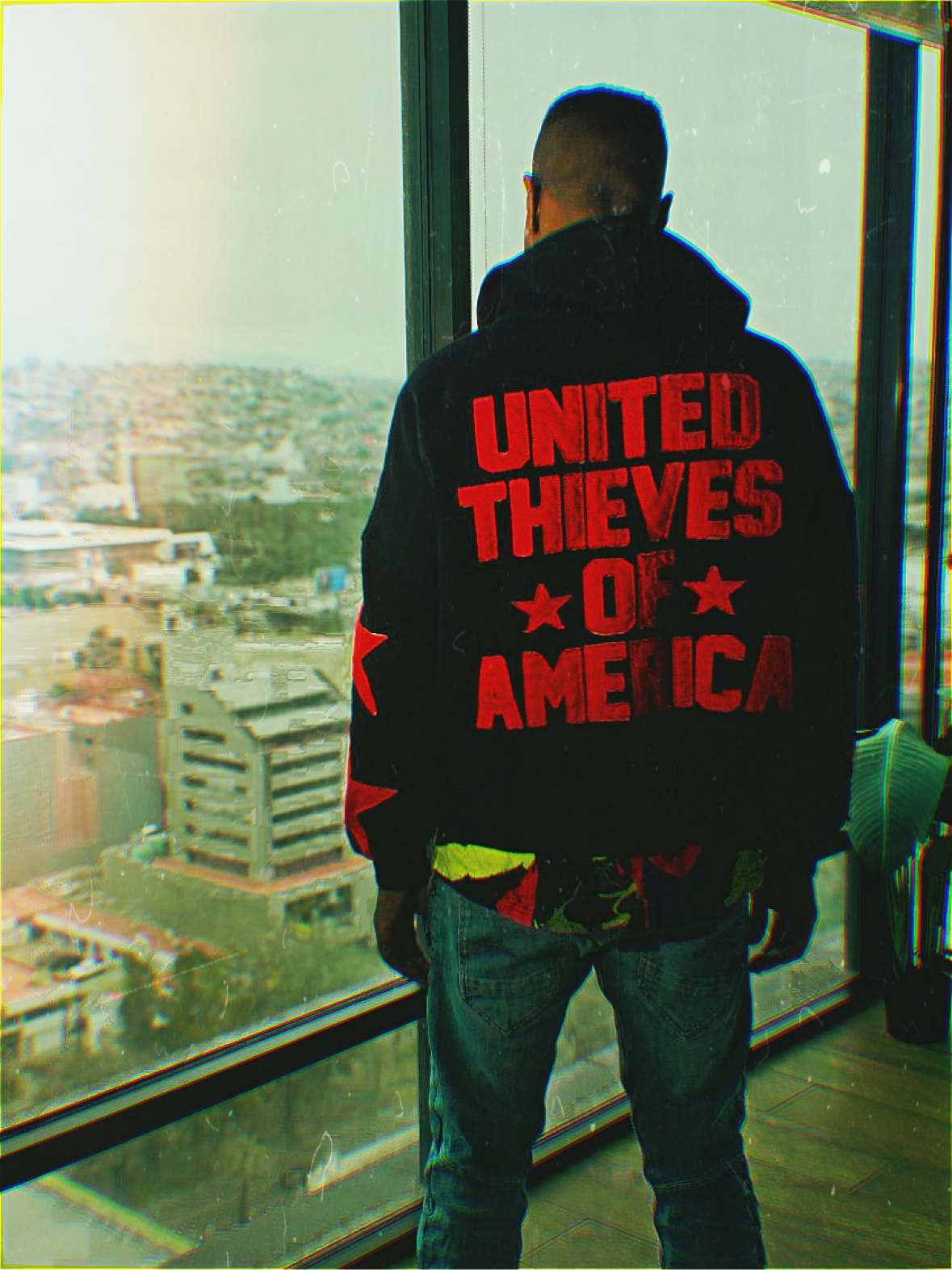 Cortez Devinci seen from behind in a black hoodie reading “UNITED THIEVES OF AMERICA” in red, standing at a high-rise window overlooking the city